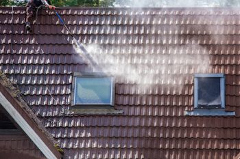 Man Worker Washing Tilted Stone Roof Shingles With Water Pressure Washer To Remove Dirt, Moss And Lichen Outdoors In Summer. Roof Cleaning Service Concept.